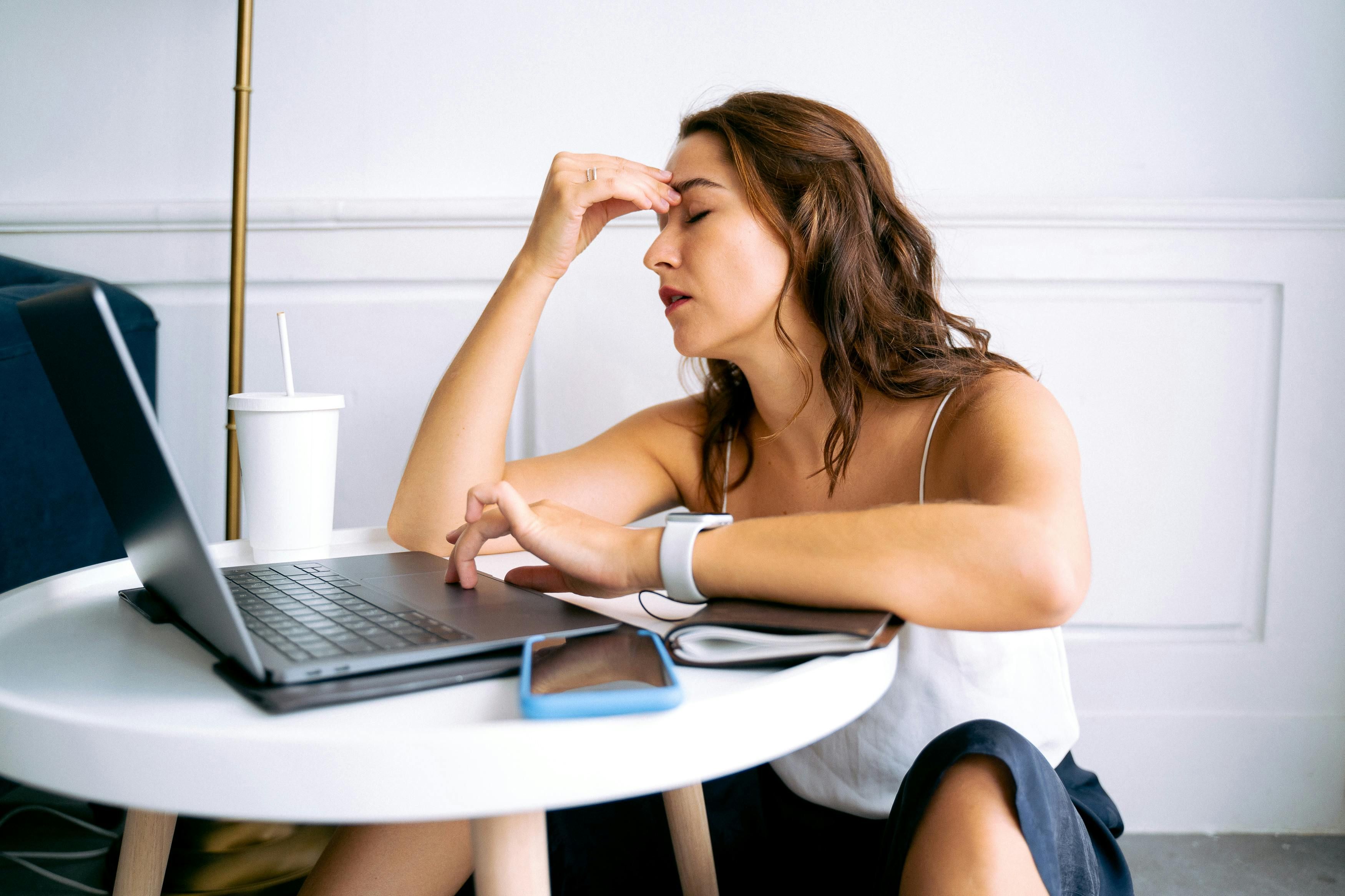 Woman Suffering from a Hormonal Migraine: Impact of Sleep Disturbances on Migraine Triggers A woman sitting in front of her computer, holding her head due to a painful hormonal migraine triggered by poor sleep.